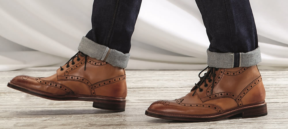 Tampilan bagian pinggir sepatu kulit berwarna cokelat, sedang dikenakan oleh seorang pria yang memakai celana jeans.